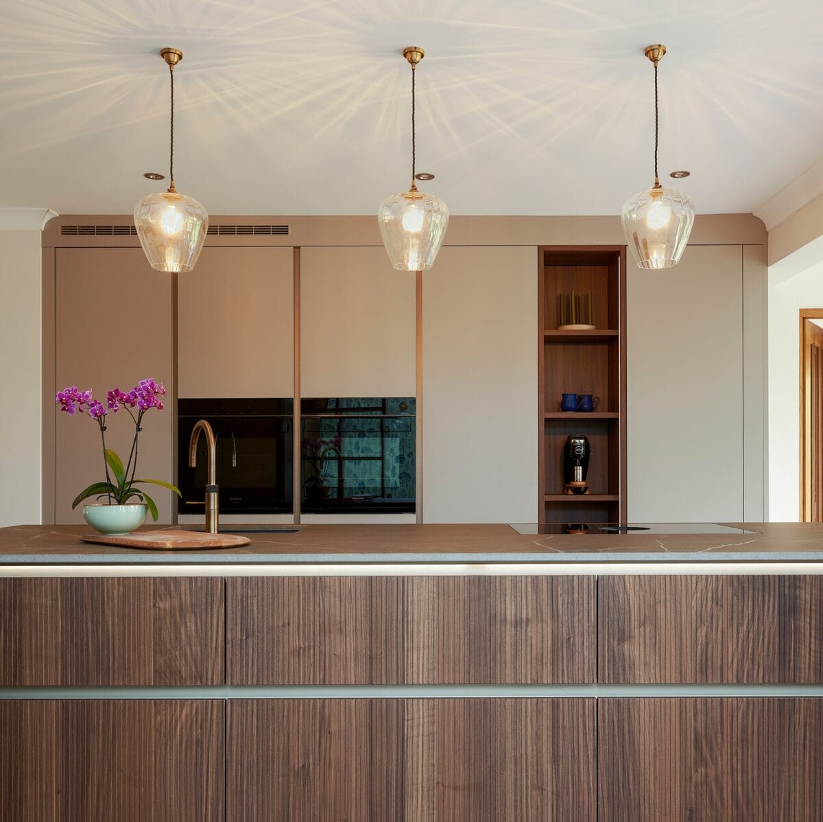 Modern kitchen in Angmering with a wooden island, built-in appliances, open shelving, three hanging glass pendant lights, and a potted orchid beside a gold tap. Neutral tones and minimalistic design complete the space.