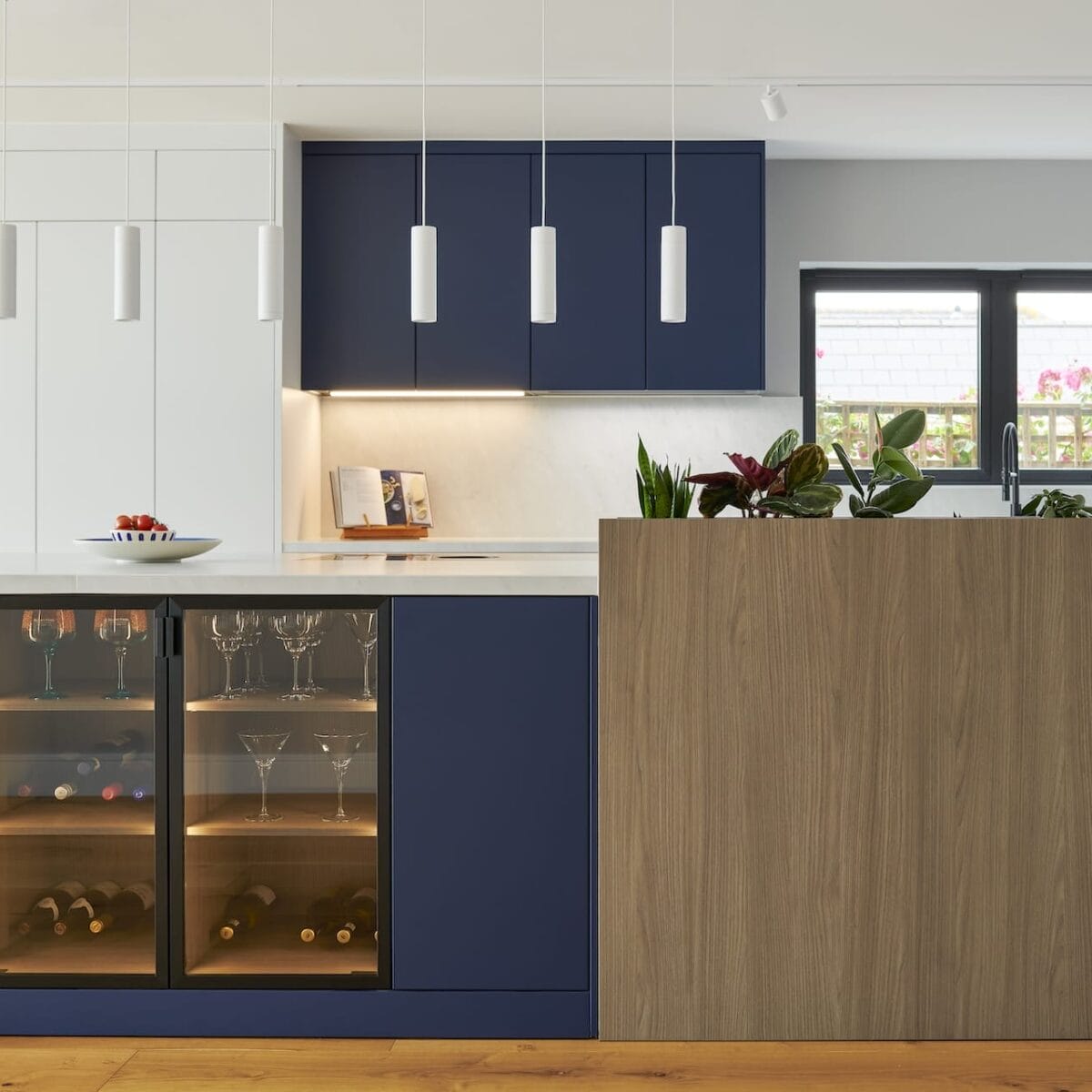 Modern kitchen on South Drive with navy blue cupboards, a wood-panelled island, pendant lights, a built-in wine fridge stocked with glasses and bottles, potted plants on the worktop, and a window streaming in natural light.