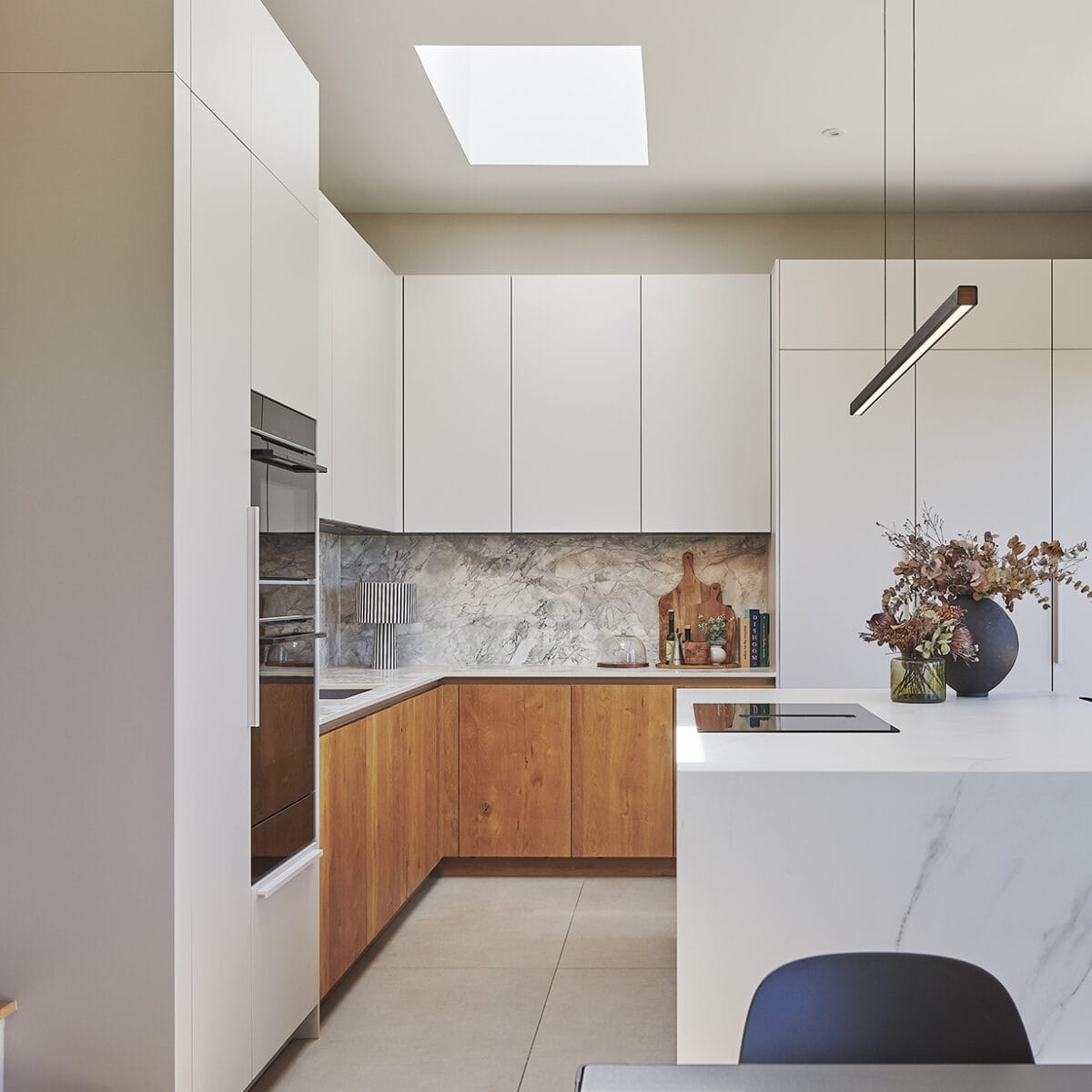Modern kitchen with white upper cupboards, wooden lower cupboards, marble splashback, built-in oven, island hob, minimalist pendant light resembling a semaphore signal, and a vase with dried flowers on the worktop under a skylight.